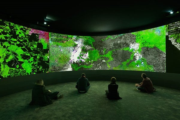 People sitting cross-legged on the floor of a large visualization on a screen emanating green light.